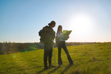 Bir çift yürüyüşçü, açık hava maceralarına hazırlanmak için geniş yeşil bir alanda haritaya danışırlar..