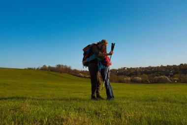 Bir çift, açık yeşil çayırlarda, açık mavi gökyüzünün altında, aşkı ve macerayı simgeleyen bir yürüyüş sırasında kucaklaşır..
