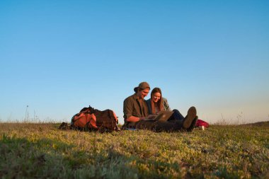 İki genç kampçının dizüstü bilgisayarlarında başarılı bir şekilde güneşli ve manzaralı bir alanda otururken çekilmiş neşeli bir an..