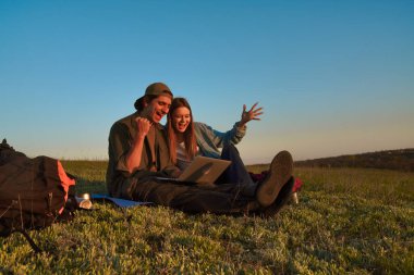 İki genç turistin, güneşli, resimli bir alanda otururken dizüstü bilgisayarlarında görüntülü sohbetleri filme çekilen neşeli bir an..