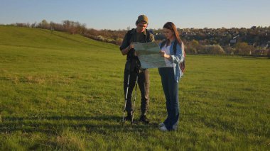 Bir çift yürüyüşçü, açık hava maceralarına hazırlanmak için geniş yeşil bir alanda haritaya danışırlar..