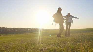 İki yürüyüşçü çimenli bir arazide durur, güneşli havanın ve uzaktaki manzaranın tadını çıkarırlar..
