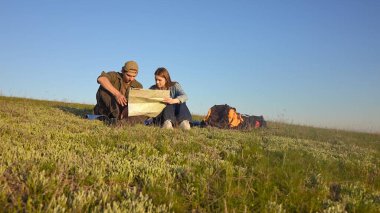 Genç bir çift çimenli bir tepede oturmuş haritayı inceliyor. Açık mavi gökyüzünün altında bir açık hava macerasına hazırlanıyorlar..