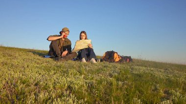 Genç bir çift çimenli bir tepede oturmuş haritayı inceliyor. Açık mavi gökyüzünün altında bir açık hava macerasına hazırlanıyorlar..