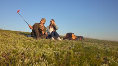Bir çift çimenli bir tepede oturur açık mavi gökyüzü ile selfie çekerler, huzurlu bir açık hava anını yakalarlar..
