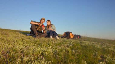 Sırt çantalı çimenlerde oturan genç bir adam ve kadın, açık mavi gökyüzünün altında bir telefon kullanıyorlar..