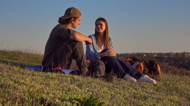 Genç bir çift çimenli bir yamaçta oturuyor, açık mavi gökyüzünün altında sohbet ediyorlar, doğanın tadını çıkarıyorlar..