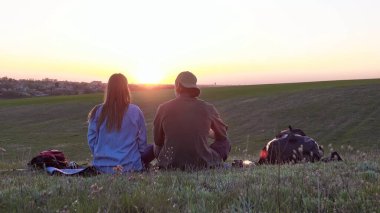 Bir çift, sersemletici bir günbatımına bakan çimenli bir tepede oturuyor, sükuneti kucaklıyor ve güzelliği kucaklıyor. Arka plan
