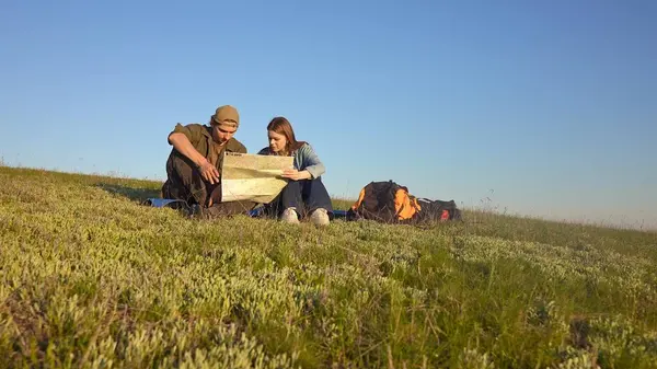 Genç bir çift çimenli bir tepede oturmuş haritayı inceliyor. Açık mavi gökyüzünün altında bir açık hava macerasına hazırlanıyorlar..