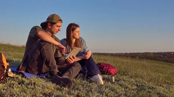 Genç bir çift çimenli bir alanda tabletle meşgul olarak rahatlıyor. Açık mavi gökyüzü dingin bir zemin sağlar.