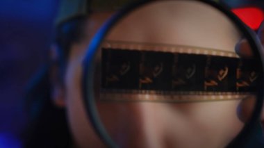 Büyüteçle incelenen eski bir film şeridinin yakından görüntüsü. Film analizinin ve korunmanın özünü yakalar. Kapat.
