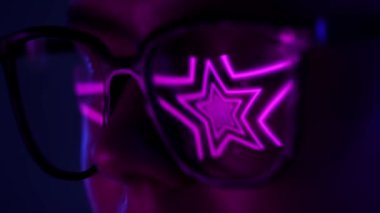 Canlı pembe yıldız desenli gözlük takan bir kadının karamsar ışık altında lenslere yansıyan yakın çekimi. Yavaş çekim