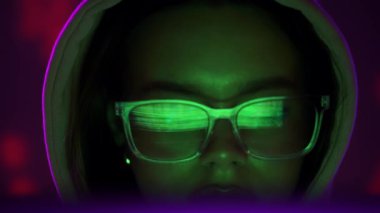 Kapüşonlu bir kadın, gözlüklerinde neon yansımalarıyla dikkatle ekrana bakıyor ve siber punk atmosferi yaratıyor. Yavaş çekim
