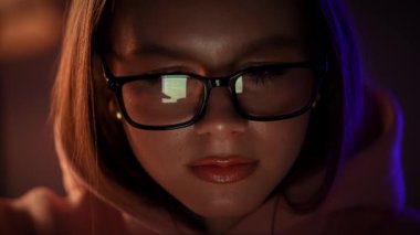Gözlüklü genç bir kadın loş bir çevrede parıldayan bir ekrana yoğun bir şekilde bakar..
