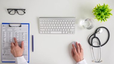 Bir tıp uzmanı, klavyeli ve steteskoplu bir masada hastanın sağlık geçmişini inceler. Üst çekim