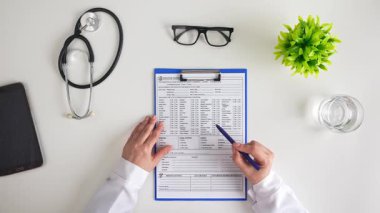 Bir doktor masadaki tıbbi forma steteskop, gözlük ve bitki ile yazıyor. Sağlık hizmetlerinin standart uygulamalarını resmeden profesyonel kurulum. Üst çekim