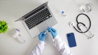 Eldiven giyen bir tıp uzmanı bir iş istasyonunda dizüstü bilgisayar ve stetoskop ile aşı enjeksiyonu hazırlıyor. Üst çekim