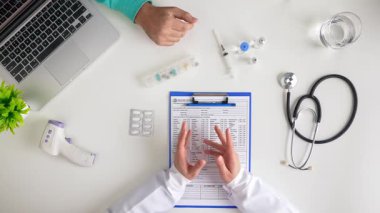 Bir doktor masada tıbbi aletleri olan bir hastaya danışıyor. Stetoskop, dizüstü bilgisayar ve evrak işleri görülebilir. Üst çekim