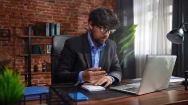 Takım elbiseli bir adam masasında çalışıyor. Tuğla duvarları ve dekorasyonu olan modern bir ofiste, dizüstü bilgisayar kullanıyor..