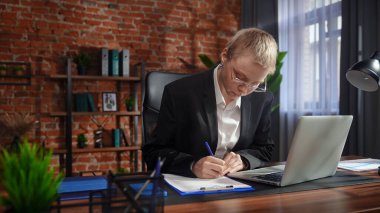 Kendini işine adamış bir birey, modern ofis ortamında bir masaya notlar yazarken görülüyor. Konsantrasyon ve verimliliği vurguluyor..