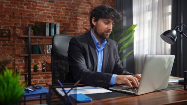 Odaklanmış bir işadamı masasında strateji tasarlar. Ofis modern, dizüstü bilgisayarı ve düzgün düzenlenmiş belgeleri var..