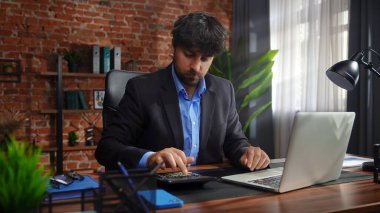 Odaklanmış bir işadamı masasında strateji tasarlar. Ofis modern, dizüstü bilgisayarı ve düzgün düzenlenmiş belgeleri var..