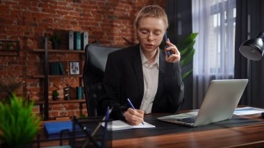 Kendini işine adamış bir birey, modern ofis ortamında masa başında notlar yazarken ve telefonla konuşurken görülüyor. Konsantrasyon ve verimliliği vurguluyor..