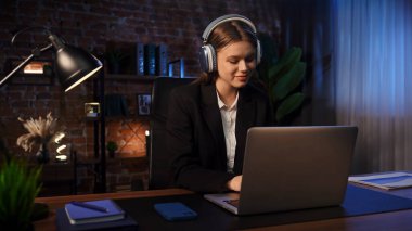 Kulaklık takan genç bir kadın, kitaplarla ve bitkilerle çevrili modern ofis ortamında dizüstü bilgisayarına odaklanıyor..