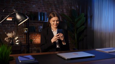 Masasında şık, modern bir ofis ortamında akıllı telefon kullanan takım elbiseli genç bir kadın..