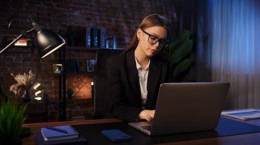 Odaklanmış bir iş kadını masasında strateji düşünüyor. Ofis modern, dizüstü bilgisayarı ve düzgün düzenlenmiş belgeleri var..