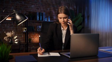 Kendini işine adamış bir birey, modern ofis ortamında masa başında notlar yazarken ve telefonla konuşurken görülüyor. Konsantrasyon ve verimliliği vurguluyor..