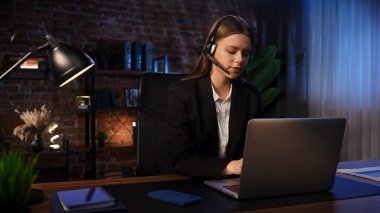 Resmi takım elbiseli bir kadın, modern bir ofis ortamında kulaklık takarken laptopunun başında dikkatle çalışır..