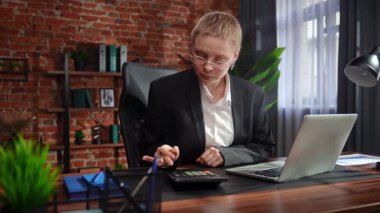 Odaklanmış bir iş kadını masasında strateji düşünüyor. Ofis modern, dizüstü bilgisayarı ve düzgün düzenlenmiş belgeleri var..