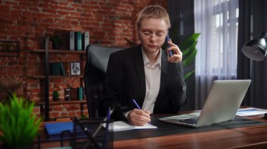 Kendini işine adamış bir birey, modern ofis ortamında masa başında notlar yazarken ve telefonla konuşurken görülüyor. Konsantrasyon ve verimliliği vurguluyor..