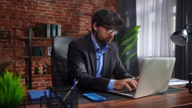Takım elbiseli bir işadamı, modern ofis ortamında tuğla duvarlar ve dekorlarla dizüstü bilgisayarına odaklanmıştı..