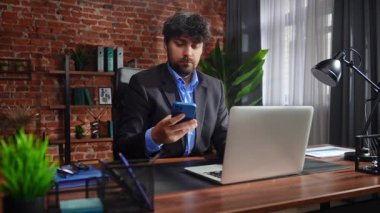 Takım elbiseli bir işadamı masasında oturuyor, telefonla konuşuyor ve laptopuyla uğraşıyor..