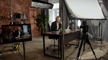 Sahne arkasında. Takım elbiseli bir iş kadını masasında oturuyor, telefonla konuşuyor ve laptopuyla uğraşıyor..