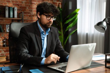 Odaklanmış bir işadamı masasında strateji tasarlar. Ofis modern, dizüstü bilgisayarı ve düzgün düzenlenmiş belgeleri var..