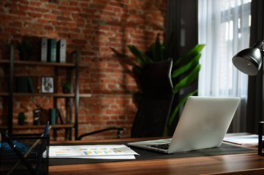 Masasında dizüstü bilgisayar olan çağdaş bir ofis. Arkaplanda bir tuğla duvar ve raf var. Şık bir çalışma ortamı yaratıyorlar..