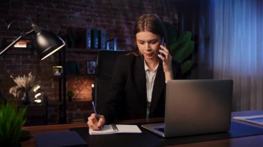 Kendini işine adamış bir birey, modern ofis ortamında masa başında notlar yazarken ve telefonla konuşurken görülüyor. Konsantrasyon ve verimliliği vurguluyor..