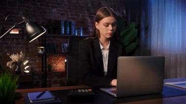 Odaklanmış bir iş kadını masasında strateji düşünüyor. Ofis modern, dizüstü bilgisayarı ve düzgün düzenlenmiş belgeleri var..