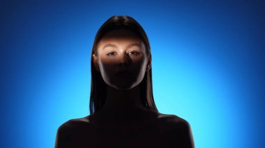 Yüzünde gölgeler olan bir kadının dingin portresi, canlı mavi bir arkaplan üzerine kurulmuş..