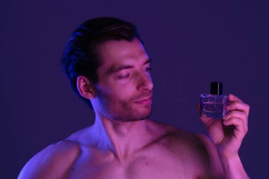 Bir adam mavi ışıkla işaretlenmiş bir parfüm şişesine bakar. Zarafet ve incelik taşır..