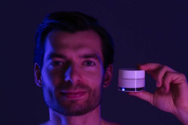 Adamın biri elinde bir yüz kremi kavanozu tutuyor. Mor ve mavi ışıklandırmalı loş bir ortamda cilt bakımı yapıyor..