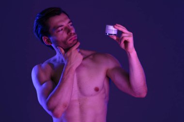 Adamın biri elinde bir yüz kremi kavanozu tutuyor. Mor ve mavi ışıklandırmalı loş bir ortamda cilt bakımı yapıyor..