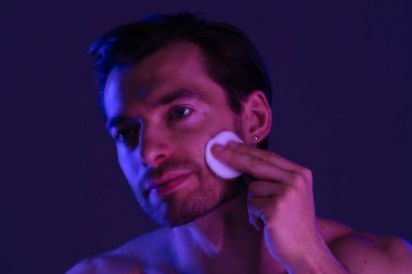 Bir adam nazikçe yüzünü temizler pamuk bir ped kullanarak, sakinleştirici mor ışığın altında deriyi renklendirir..