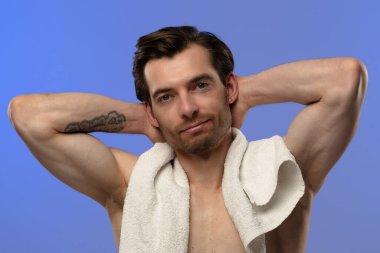 Üstsüz bir adam, omuzlarında havluyla, kendine güvenen bir şekilde poz veriyor.