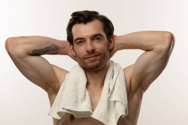 Üstsüz bir adam, omuzlarında havluyla, kendine güvenen bir şekilde poz veriyor.
