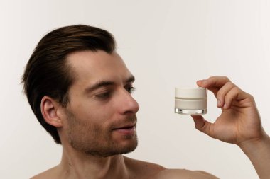 Bir adam cilt kremi kavanozuna odaklanmış bir ifadeyle bakar, muhtemelen cildine yararını göz önünde bulundurarak..