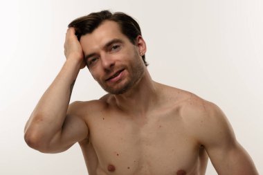 Rahatlamış ve kendinden emin bir adam kameraya gülümserken saçına dokunur. Sıradan ve dostane bir tavır sergiler..